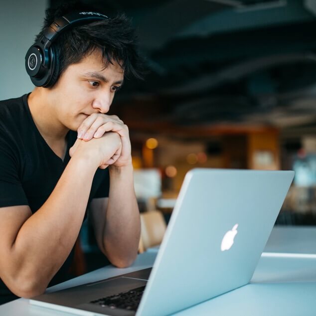 A man with headphones on, seated at table looking at his laptop. 