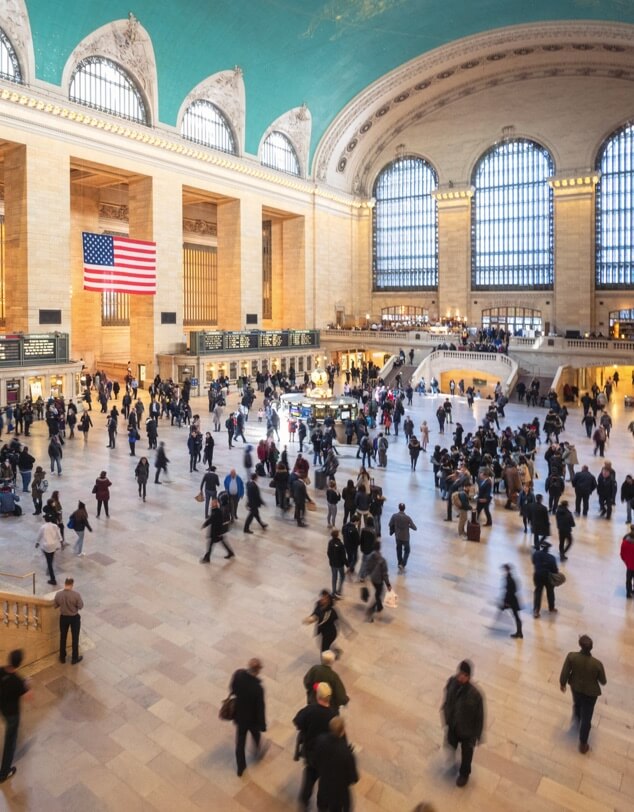 A large group of businesspeople walking across a train station beneath an American flag. 