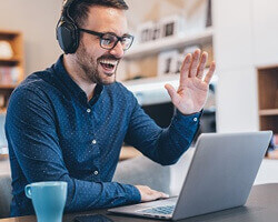 A smiling Caucasian or Latino man working on a laptop while wearing headphones, raising his hand. 