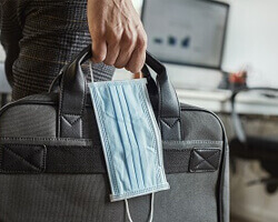 A person's hand holding a briefcase and a surgical mask. 